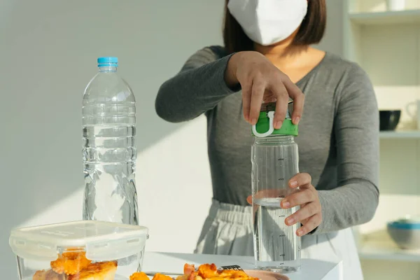 Woman wearing white mask pouring water from bottle in kitchen.