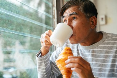 Yaşlı adam elinde sosisli kruvasan tutarken bardaktaki sıcak kahveyi içerken camda hoparlör açık, çocuğunu dışarıda onu ziyaret ederken görünce şaşırdı..