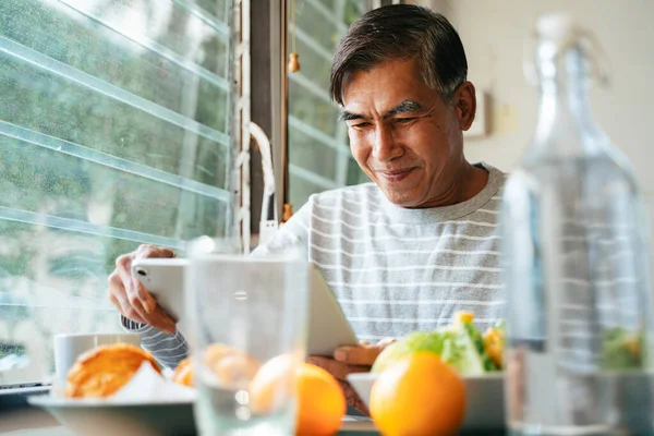 Old man with gray hair sitting at the table in kitchen with breakfast on the table, he reading the news in tablet and smile about funny news.