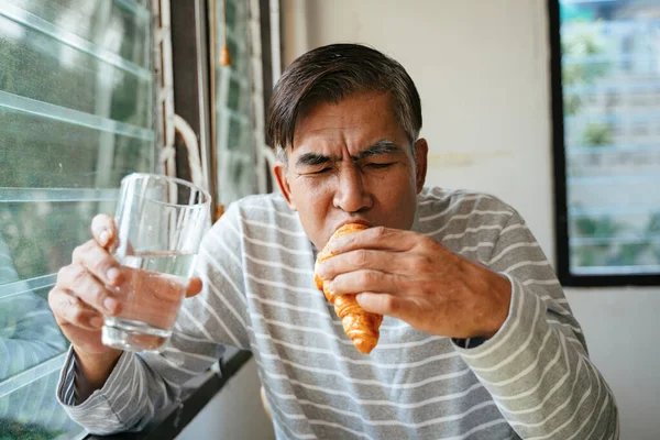 Old man eating sausage croissant while hold a glass of drinking water in another hand.