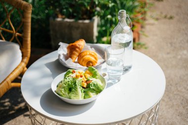 Salata ve sosisli kruvasan da temiz şişe içinde içme suyu ve güneş ışığında yuvarlak masanın üzerindeki cam..
