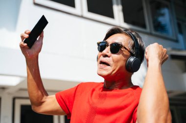 Kırmızı tişörtlü yaşlı adam güneşli bir günde evinde mobil uygulama müziğinin keyfini çıkarıyor..