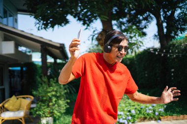 Kırmızı tişörtlü yaşlı adam akıllı telefonu tut ve gerçek kablosuz kulaklığı tak ve evinde güneş ışığında dans et..