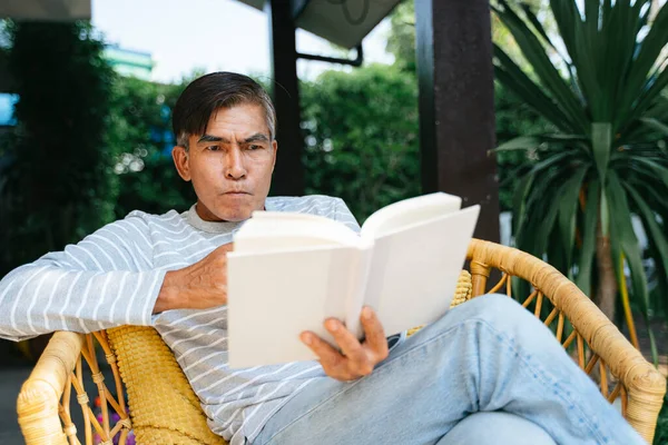 Old man reading the book and srink the water while sitting on the wicker bench in the yard.