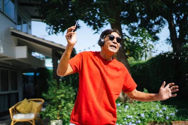 Old man in red t-shirt hold the smartphone and wearing the true wireless headphone and dance in the sunlight at his home.