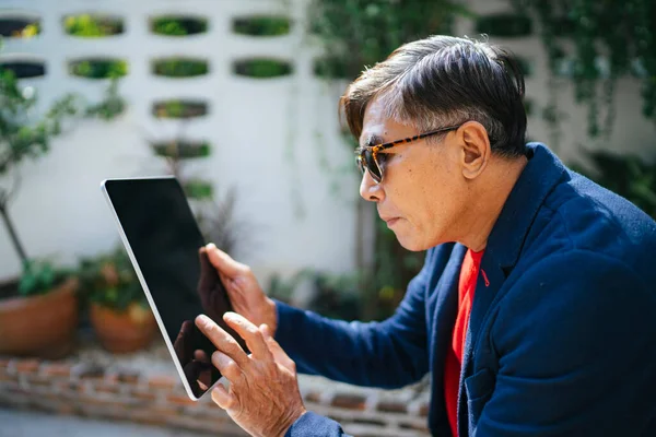 Old man wearing blue suit and eyeglasses attentively watching the information about stock market with tablet and focus on the information.