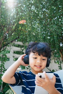 Kablosuz kulaklık takan küçük çocuk hafta sonu arka bahçede oturup akıllı telefondan çizgi filmi izliyor..