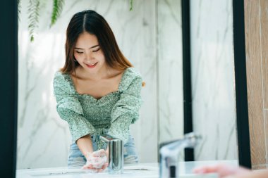 Beautiful asian woman on fashion green shirt washing hands with water and soap bubble in toilet bathroom. Looking at the mirror. clipart