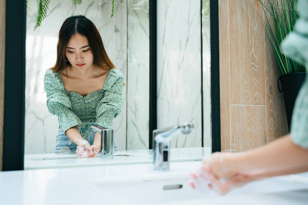 Beautiful asian woman on fashion green shirt washing hands with water and soap bubble in toilet bathroom. Looking at the mirror.