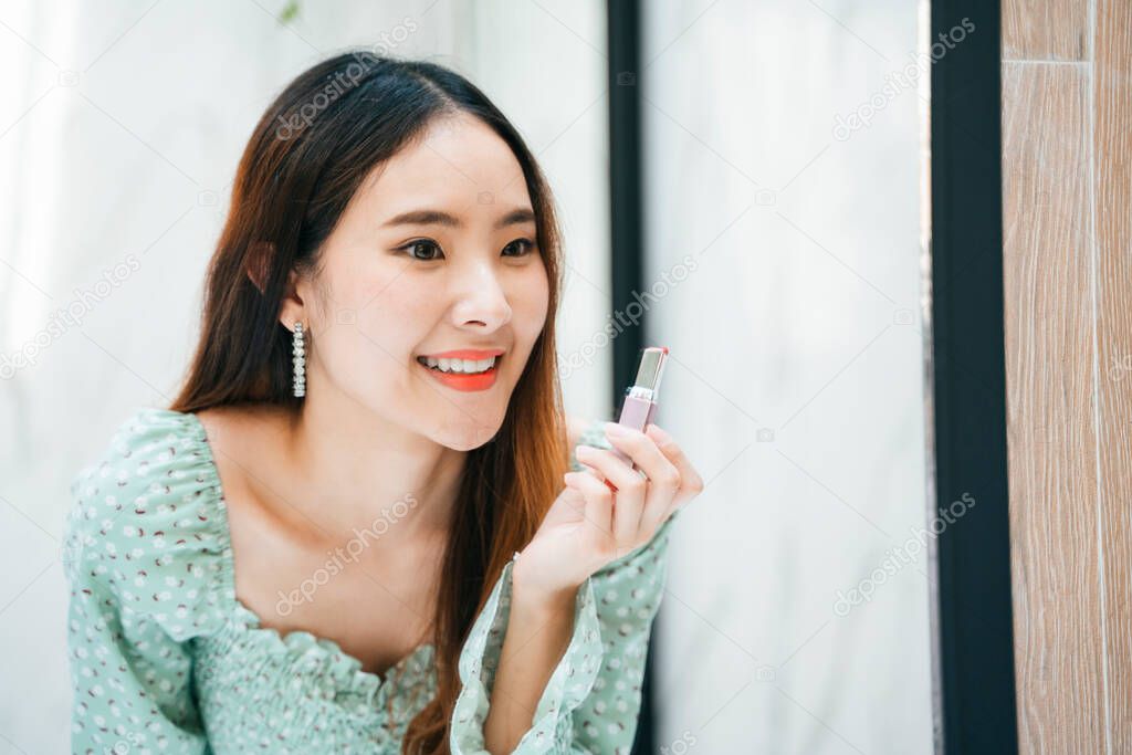 Cheerful beautiful young asian woman on green fashion shirt applying lipstick in front of the mirror.