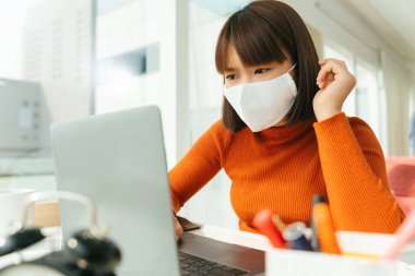 Genç, güzel Asyalı Taylandlı kısa saçlı, beyaz cerrahi maske takan, laptopla evde ders çalışan bir kadın..