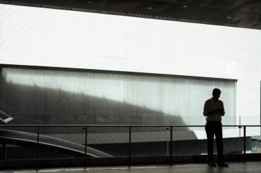 Modern ofisin dışında akıllı telefon kullanan beyaz tişörtlü Asyalı bir iş adamının silüeti. Büyük beyaz banknot tahtası.