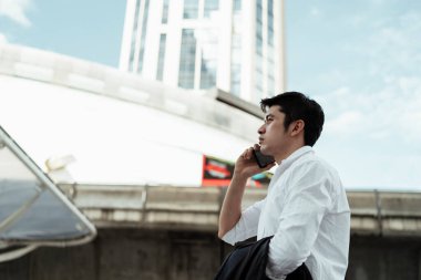 Beyaz tişörtlü, akıllı telefon kullanan, büyük şehirde telefonla konuşan olgun Asyalı iş adamı..