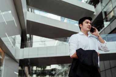 Beyaz tişörtlü Asyalı bir iş adamı sokakta akıllı telefondan konuşuyor..