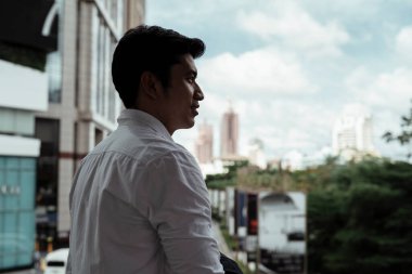Beyaz tişörtlü, olgun Asyalı iş adamı balkonda dikilip şehre bakıyor..