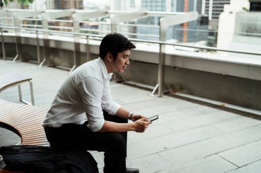 Beyaz tişörtlü olgun bir Asyalı iş adamı çatıdaki bankta oturup akıllı telefon çalmayı sever..