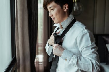 Beyaz tişörtlü Asyalı Taylandlı adam evde boynuna kravat takıyor. Boyun kravatı giydirme prosedürü.