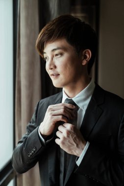 Blac kostümlü Asyalı adam işe gitmeden önce boyun kravatını düzeltiyor. Şatafatlı ve zeki bir adam odayı süslüyor..