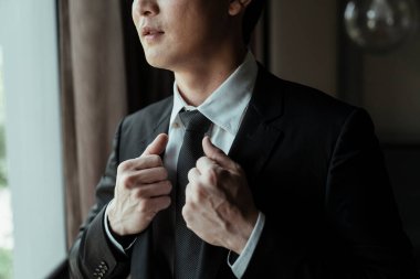 Blac kostümlü Asyalı adam işe gitmeden önce boyun kravatını düzeltiyor. Şatafatlı ve zeki bir adam odayı süslüyor..