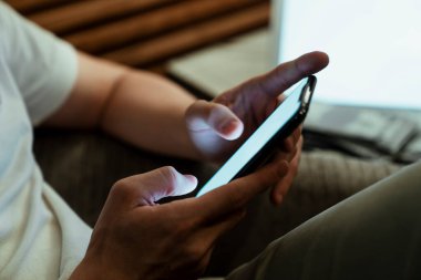 Erkek ellerinin arka planda dizüstü bilgisayarında beyaz boşluk olan akıllı telefonu kullandığının yakın plan görüntüsü. Koltukta oturan ve cep telefonu kullanan bir adam..