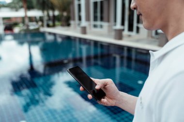 Elinde akıllı telefon tutan Asyalı adam havuzun kenarında yürüyor..