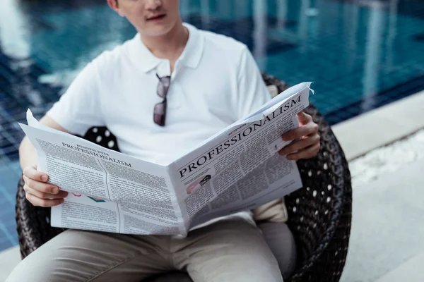 Asian rich guy businessman reading news paper at resort sitting on a chair near swimming pool.