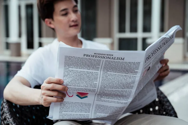 Asian rich guy businessman reading news paper at resort sitting on a chair near swimming pool.