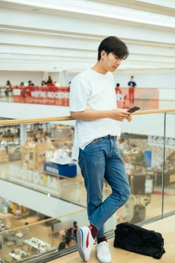 Kot pantolonlu ve beyaz tişörtlü genç adam asma katta akıllı telefonuyla dururken barikata yaslanıyor..