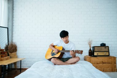 Minimum yatak odasında beyaz yatakta oturan siyah saçlı müzisyen gitar çalıyor..