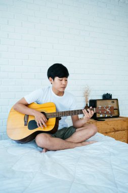 Minimum yatak odasında beyaz yatakta oturan siyah saçlı müzisyen gitarını dikey çalıyor..