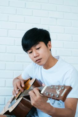 Beyaz odada oturan siyah saçlı müzisyen gitar çalıyor..