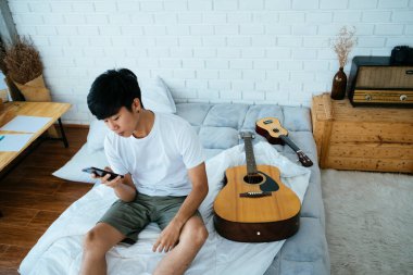 Siyah saçlı müzisyen beyaz yatakta gitar ve ukulele ile oturuyor ve akıllı telefonu kullanıyor..