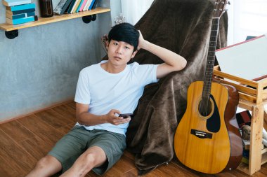 Siyah saçlı müzisyen, tahta zeminde oturuyor kahverengi sandalyeye yaslanıyor ve akıllı telefonu tutuyor..