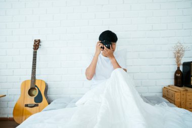 Siyah saçlı adam beyaz yatakta oturuyor ve kameraya bakmak için fotoğraf çekiyor..