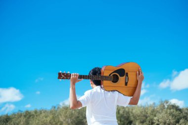 Mavi gökyüzünün altında ormanda dururken siyah saçlı adam gitarı omuzunda taşıyor..