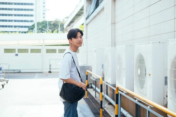 Teenage guy in jeans and white t-shirt standing at the shadow of white building with black bag.