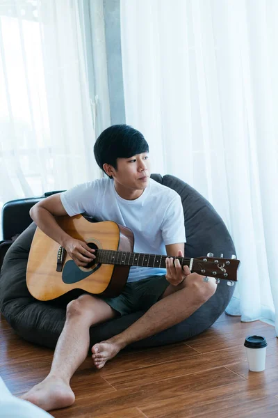 Black hair musician guy sitting on black beanbag and playing guitar with a cup of coffee on the wooden floor.