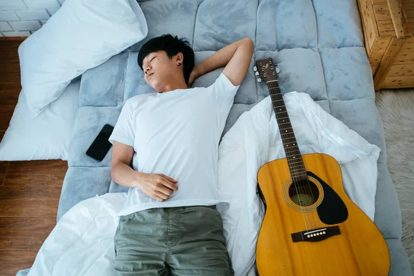 黒い髪のミュージシャン男は ギターとスマートフォンで白いベッドの上で眠る — ストック写真