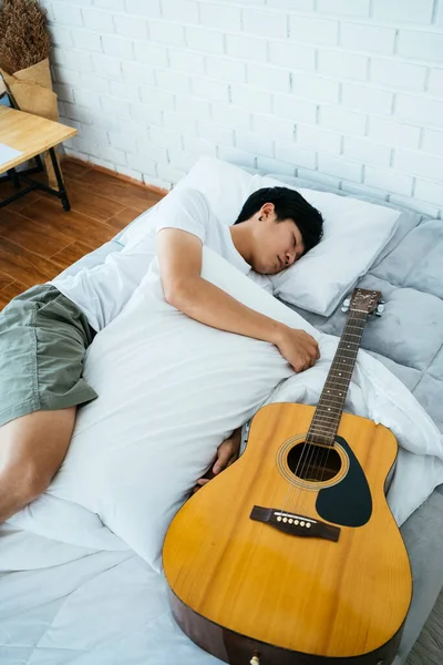 Black hair musician guy sleep on the white bed with guitar and hug the pillow.