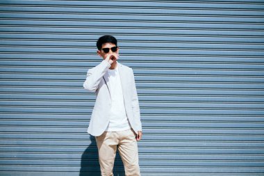 Beyaz takım elbiseli bir adam güneş gözlüğü takıyor. Fabrikanın metal kepenk kapısının önünde duruyor..