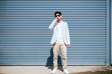 Beyaz takım elbiseli bir adam güneş gözlüğü takıyor. Fabrikanın metal kepenk kapısının önünde duruyor..