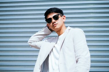 Beyaz takım elbiseli bir adam güneş gözlüğü takıyor. Fabrikanın metal kepenk kapısının önünde duruyor..