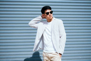 Beyaz takım elbiseli bir adam güneş gözlüğü takıyor. Fabrikanın metal kepenk kapısının önünde duruyor..