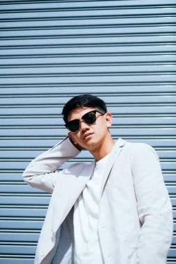 Beyaz takım elbiseli bir adam güneş gözlüğü takıyor. Fabrikanın metal kepenk kapısının önünde duruyor..