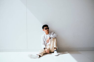 Beyaz takım elbiseli, güneş gözlüklü bir adam beyaz duvarda ışık ve gölgeli bir model gibi davranır..