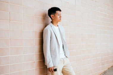 Beyaz takım elbiseli bir adam tuğla duvarda duruyor. Elinde güneş gözlüğü var..