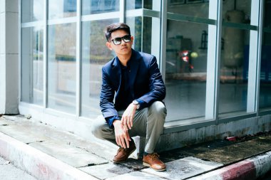 Koyu mavi takım elbiseli, güneş gözlüklü bir adam güneş ışığında binanın patikasında oturuyor..