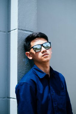 Koyu mavi gömlekli, güneş gözlüklü bir adam güneş ışığında binaya yaslanıyor..