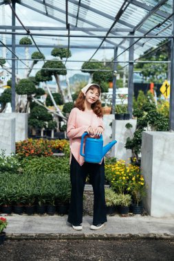 Bej saç bandı ve pembe uzun kollu tişörtlü esmer kız güneşli bir günde cam evde elinde kırık su kabıyla dikiliyor..