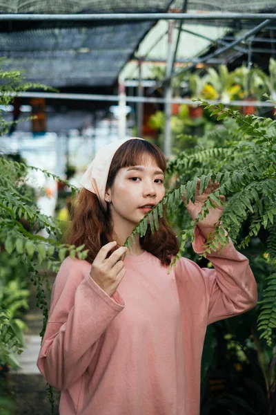 穿着米色头巾和粉色长袖T恤的布鲁内特女孩站在温室里挑选蕨类 — 图库照片